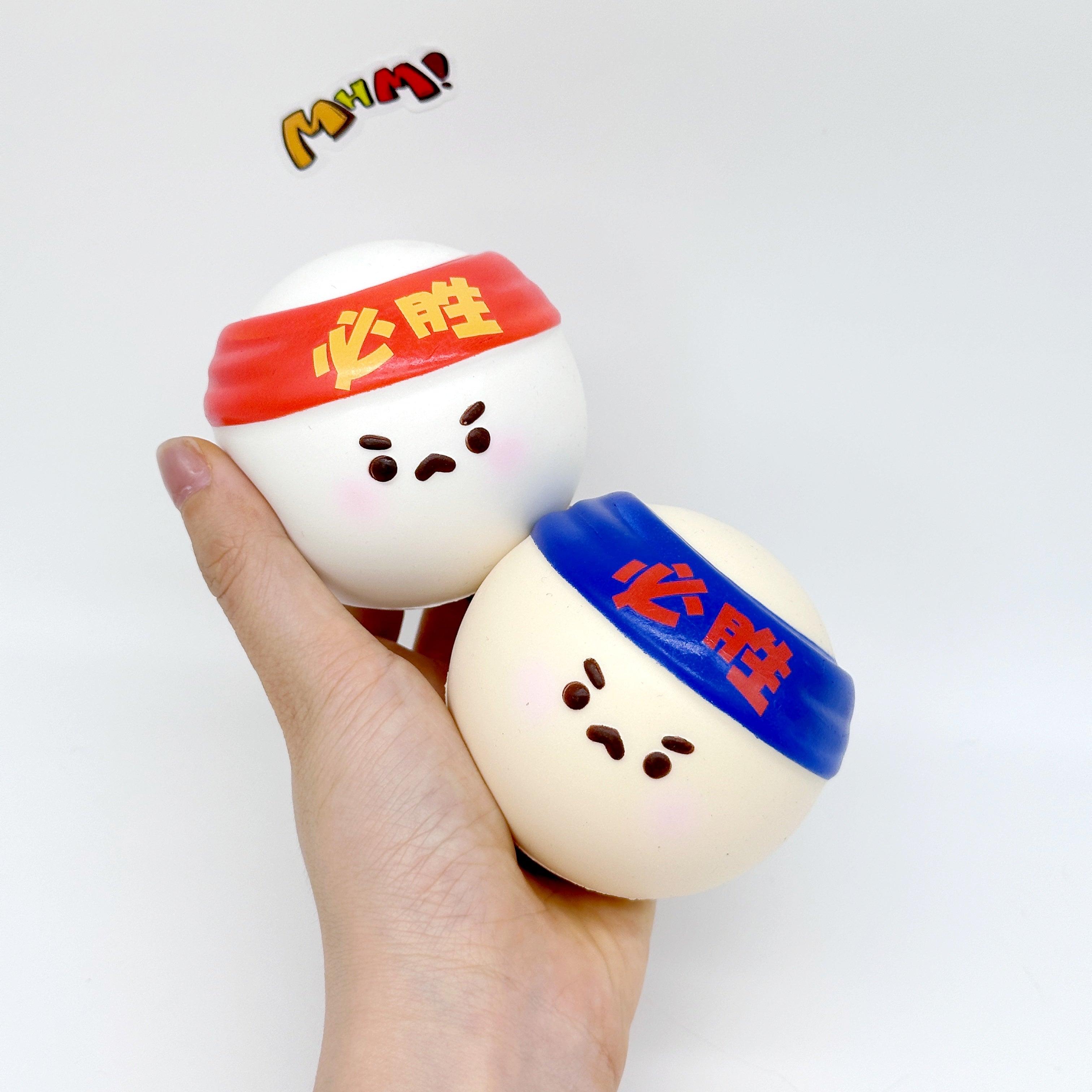 Hand holding two Must Win ping pong ball squishy toys with cute faces and red blue headbands
