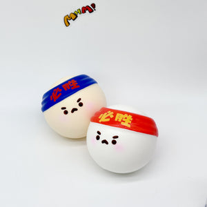 Two white ping pong ball squishies with blue and red Must Win headbands, angry faces, side by side on white background